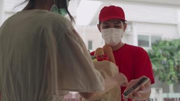 der lieferbote trägt rote hemden und masken, die lebensmitteltütenpapier halten. Die Mitarbeiter stehen vor der Haustür der Kunden, die Produkte online bestellen. Lebensmittelbote klingelte für gesendete Waren. video