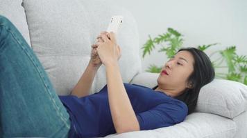 bela jovem asiática com expressão facial deprimida dorme perto no sofá têxtil cinza segurando seu telefone. mulher triste em seu quarto. conceito de bullying cibernético. video