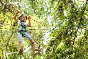 adorable niña disfrutando de su tiempo en el parque de aventuras de escalada en un cálido y soleado día de verano. actividades de verano para niños pequeños. niño divirtiéndose en vacaciones escolares. foto