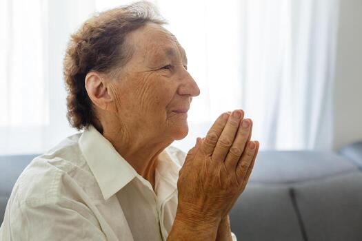 Faithful old senior grandmother pray with hope faith holding hands together sit on sofa alone at home, religious elderly woman believer christian with eyes closed say gratitude worship prayer concept photo