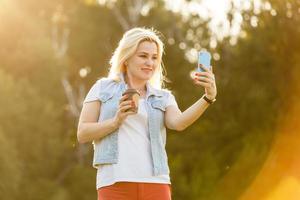 Beautiful happy young woman taking a selfie photo with smart phone outdoors in park on summer day. Modern millennial youth lifestyle and travel concept.