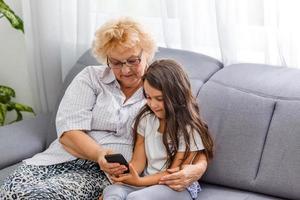 Happy grandmother and cute granddaughter using cellphone together, smiling older grandma and child girl having fun taking selfie on phone, cheerful granny with little kid play making photo on mobile