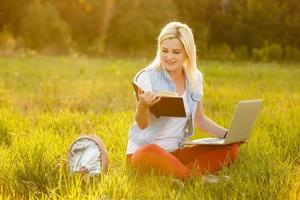 Portrait of pretty young woman sitting on green grass in park with legs crossed during summer day while using laptop for video call photo