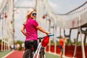 hermosa chica con cabello rubio felizmente montando en bicicleta. retrato de una joven con gafas de sol j divirtiéndose, montando en bicicleta por las calles de la ciudad foto