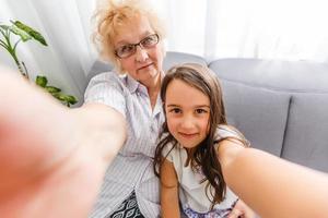 Happy grandmother and cute granddaughter using cellphone together, smiling older grandma and child girl having fun taking selfie on phone, cheerful granny with little kid play making photo on mobile