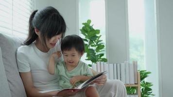 Lycklig asiatisk mor koppla av och läsa bok med bebis tid tillsammans på Hem. förälder sitta på soffa med dotter och läsning en berättelse. lära sig utveckling, barnomsorg, skrattande, utbildning, berättande, öva. video