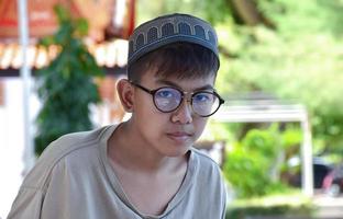 Young asian muslim boy wears eyeglasses, sitting in school park and reading his book in his free times before going back home, soft and selective focus. photo