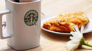 Washington, EE.UU. - 01 de agosto de 2022 taza de café con el logotipo de Starbucks en la parte delantera, panadería blanca en el plato. colóquelo en una mesa de madera en el jardín donde brilla el sol de la mañana. foto