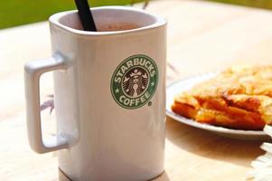WASHINGTON, USA - August 01 2022  Coffee mug with Starbucks logo on front, white bakery on plate. Place it on a wooden table in the garden where the morning sun shines. photo