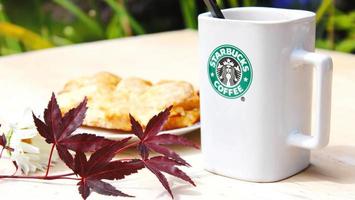 Washington, EE.UU. - 01 de agosto de 2022 taza de café con el logotipo de Starbucks en la parte delantera, panadería blanca en el plato. colóquelo en una mesa de madera en el jardín donde brilla el sol de la mañana. foto