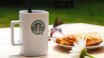 Washington, EE.UU. - 01 de agosto de 2022 taza de café con el logotipo de Starbucks en la parte delantera, panadería blanca en el plato. colóquelo en una mesa de madera en el jardín donde brilla el sol de la mañana. foto