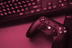 Modern gamepad lies with pc keyboard on table in dark playroom scene. Gameplay streaming and video game walkthroughts concept Image toned in Viva Magenta, color of the 2023 year photo