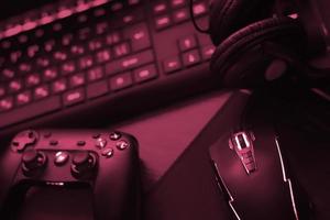 el gamepad moderno y el mouse para juegos se encuentran con el teclado y los auriculares en la mesa en la escena oscura de la sala de juegos. imagen conceptual de retos y competiciones de videojuegos tonificada en viva magenta, color del año 2023 foto