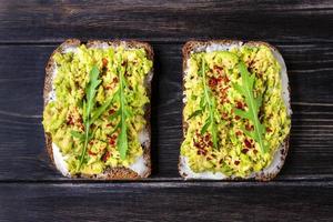 Sándwiches para el desayuno: rebanada de pan oscuro de grano entero, queso crema, guakomole, adornado con rúcula, pimentón en la mesa de madera, vista superior plana, concepto de comida saludable hecha en casa foto