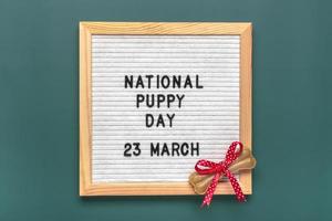 Felt board with text National puppy day in 23 march, dog accessories - bone with red bow on green background Top view Flat lay Holiday card photo