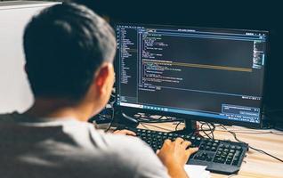 Back view young asian man sitting at desk using computer programming debugging improving binary algorithm code in workstation By focusing on the monitor and blurring on people photo