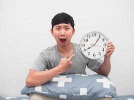 el hombre sentado en la cama se siente conmocionado apuntando con el dedo al reloj, concepto de despertarse tarde foto