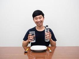 Positive man smile holding water bottle sit at table with dish and cutlery white background photo