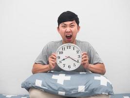 Man sit on the bed holding analog clock shocked face,Wake up late concept photo