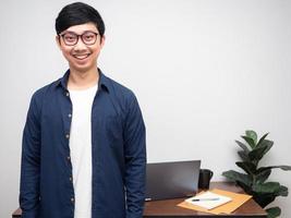 joven empresario gafas sonrisa stand frente a la mesa del lugar de trabajo en el espacio de copia de la habitación foto