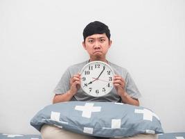 el hombre se sienta en la cama con la cara enojada del reloj analógico, despierta el concepto tarde foto