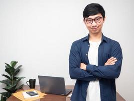 hombre de negocios positivo gafas sonrisa stand brazos cruzados confiado en la mesa de trabajo lugar de trabajo foto