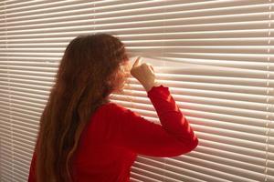 Rear view of beautiful young woman with long hair peeks through hole in the window blinds and looks out the window. Surveillance and curiosity concept photo