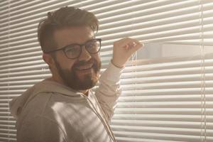 un joven con barba se asoma por un agujero en las persianas de la ventana y mira hacia la calle. concepto de vigilancia y curiosidad foto