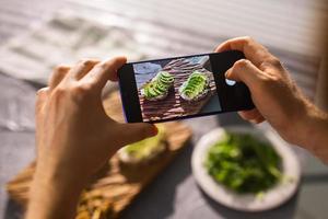 las manos toman fotos en el teléfono inteligente de dos hermosos y saludables sándwiches de crema agria y aguacate sobre la mesa. redes sociales y concepto de comida