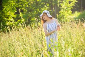 Happy young woman with long hair in hat and dress pulls her hands towards the plants while walking through the summer forest on a sunny day. Summer joy concept photo