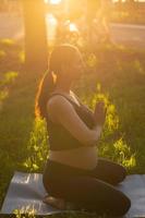 Cute young caucasian pregnant woman is meditating while sitting on a rug on the lawn on a sunny summer evening. Concept of pacification and energy boost photo