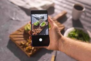 Hands take pictures on smartphone of two beautiful healthy sour cream and avocado sandwiches lying on board on the table. Social media and food concept photo