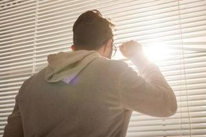 Rear view of young stylish man peeking through hole in window blinds and looking out into street. Concept of enjoying the morning sun and positivity photo