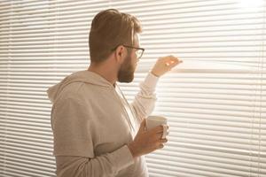 Rear view of young stylish man with beard and coffee peeking through hole in window blinds and looking out into street. Concept of enjoying the morning sun and positivity photo