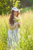 Happy young woman with long hair in hat and dress pulls her hands towards the plants while walking through the summer forest on a sunny day. Summer joy concept photo