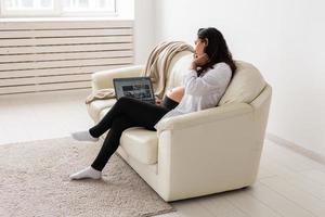 Latin pregnant woman using laptop computer sitting on sofa at home. Pregnancy and information for parenthood concept. photo