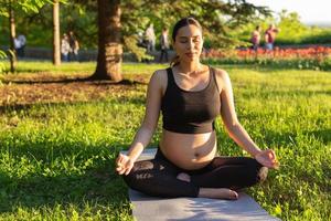 Peaceful young positive pregnant woman in gymnastic suit does yoga and meditate sitting on mat on green grass on sunny warm summer day. Concept of preparation for childbirth and positive attitude photo