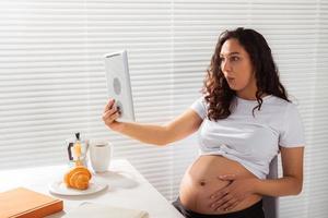 Happy pregnant young beautiful woman talking to mom using video call during morning breakfast. Concept of communication and positive attitude during pregnancy photo