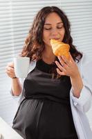 Happy pregnant young beautiful woman eating croissant during morning breakfast. Concept of pleasant morning and positive attitude during pregnancy photo