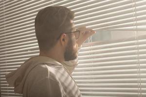Rear view of young man with beard peeks through hole in the window blinds and looks out into the street. Surveillance and curiosity concept photo