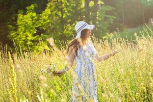 Happy young woman with long hair in hat and dress pulls her hands towards the plants while walking through the summer forest on a sunny day. Summer joy concept photo