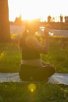 Pregnant woman drink water while sitting on yoga mat in summer park. Healthy lifestyle, expecting baby and child bearing concept. photo