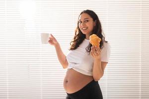 feliz embarazada joven hermosa mujer comiendo croissant durante el desayuno de la mañana. concepto de mañana agradable y actitud positiva durante el embarazo foto