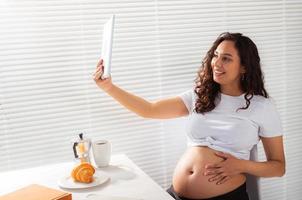 Hispanic pregnant woman using digital tablet while breakfast. Technology, pregnancy and maternity leave photo