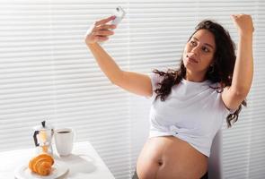 Happy pregnant young beautiful woman talking to mom using video call during morning breakfast. Concept of communication and positive attitude during pregnancy photo