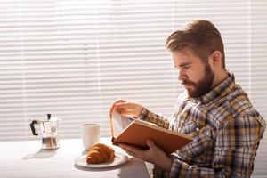 Side view of pensive young bearded hipster man reading book and having dinner with croissant and cup of coffee on background of blinds. Good morning or lunch break concept photo