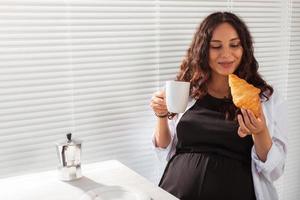 Happy pregnant young beautiful woman eating croissant during morning breakfast. Concept of pleasant morning and positive attitude during pregnancy photo