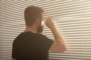 Rear view of young man with beard peeks through hole in the window blinds and looks out into the street. Surveillance and curiosity concept photo