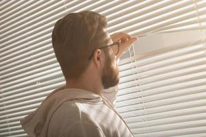 Rear view of young man with beard peeks through hole in the window blinds and looks out into the street. Surveillance and curiosity concept photo