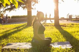 Peaceful young positive pregnant woman in gymnastic suit does yoga and meditate sitting on mat on green grass on sunny warm summer day. Concept of preparation for childbirth and positive attitude photo